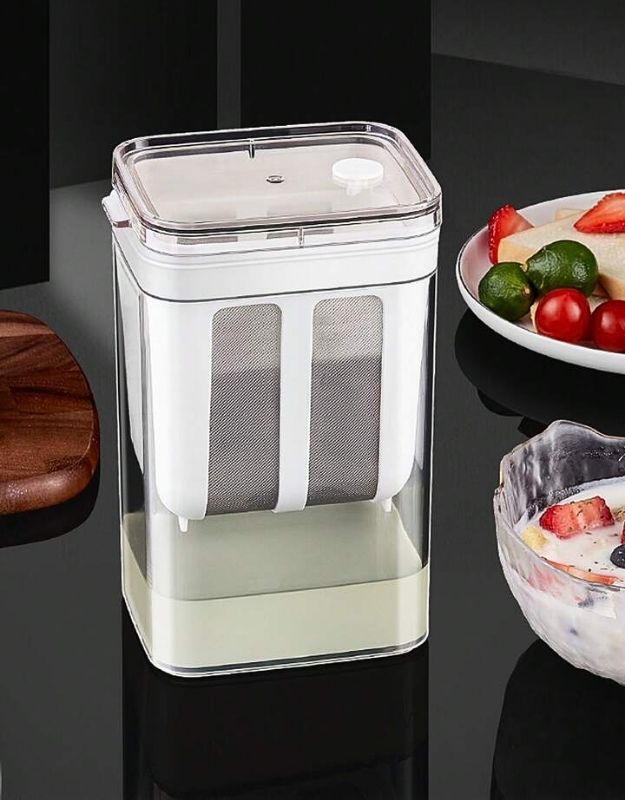 A multifunctional Greek yogurt and curd maker with a transparent lid and fine mesh strainer, displayed on a kitchen counter.
